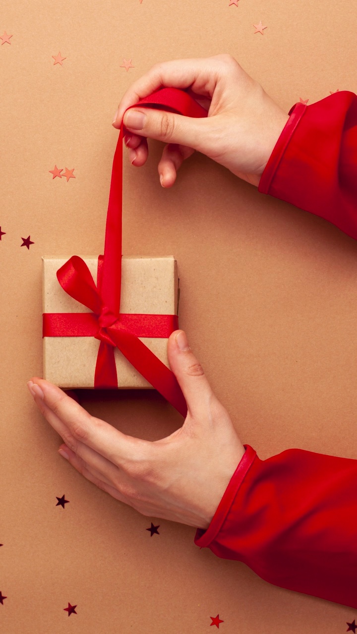 人的手用红色的袖子绑了一个棕色的纸礼物与红色缎带蝴蝶结在棕色的背景与红色的星星形状。定格动画垂直圣诞假期平铺视频下载