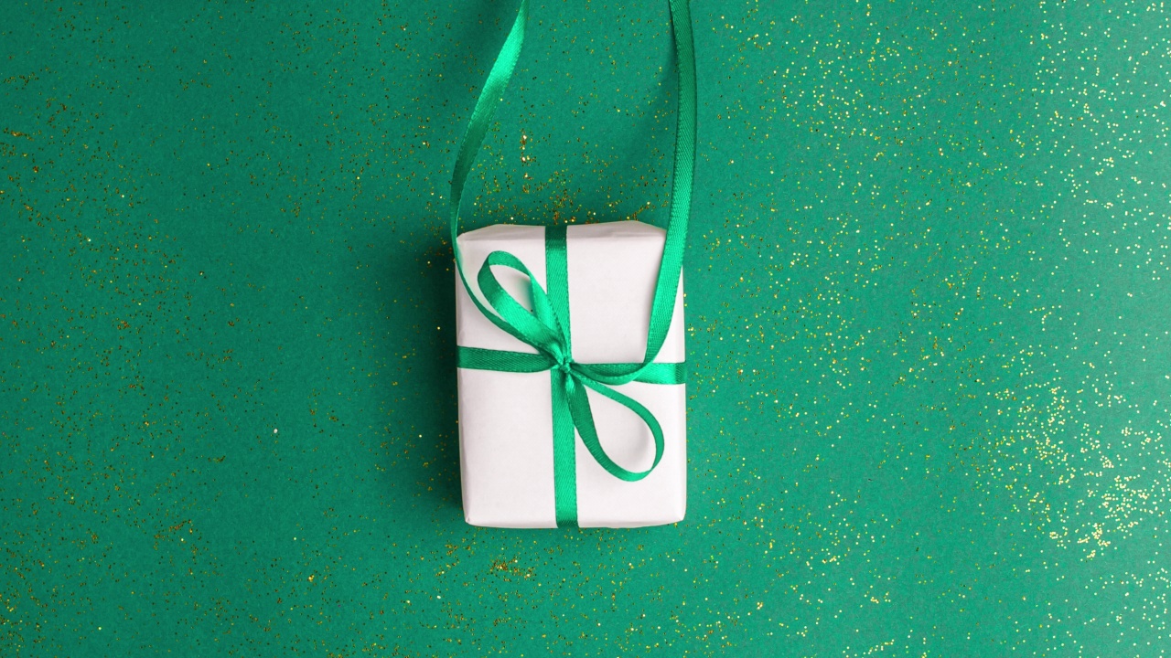 人类的手在白色的毛衣把一个白色的纸礼物翠绿色缎带蝴蝶结绿色背景与金色的闪光。定格动画圣诞假期概念平铺视频下载