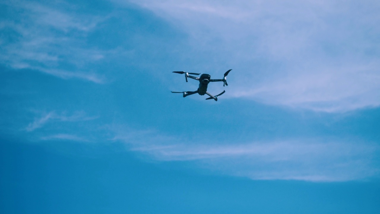 无人机在蓝天白云的背景下飞行，从下面看。装有摄像机的四轴飞行器在低空盘旋。展出无人机螺旋桨。视频下载