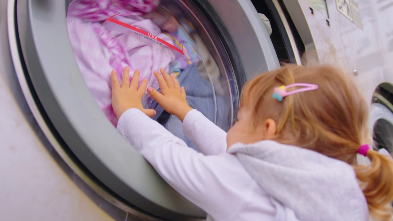 小女孩经常打扫家务，把毛巾放进洗衣机，在舒适的洗衣房里打开现代的洗衣机-烘干机。家务的概念视频下载