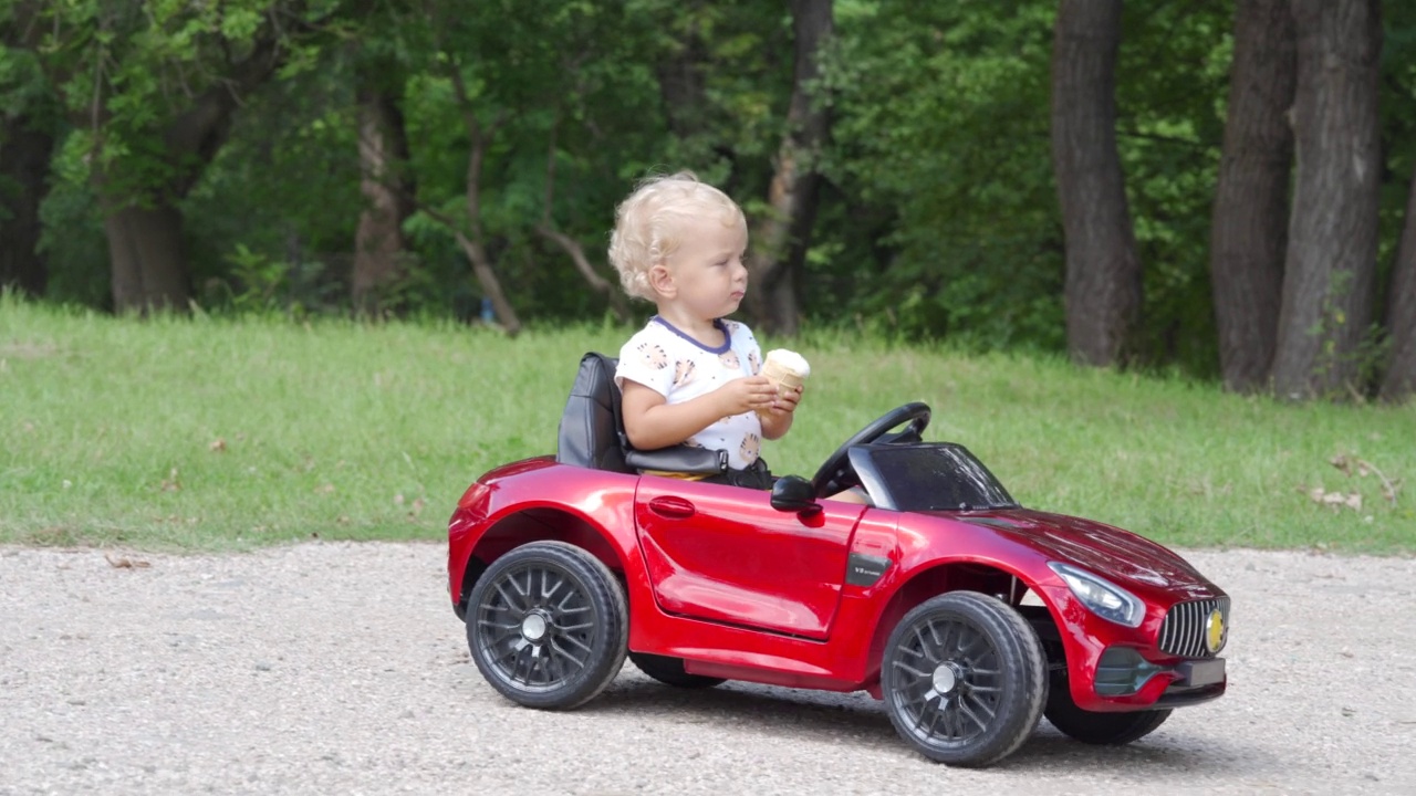 快乐的孩子在夏日公园里吃着冰淇淋，开着电动玩具车。1.5岁的幼童骑电动汽车玩得很开心。小男孩骑着小汽车玩具在户外玩。视频素材