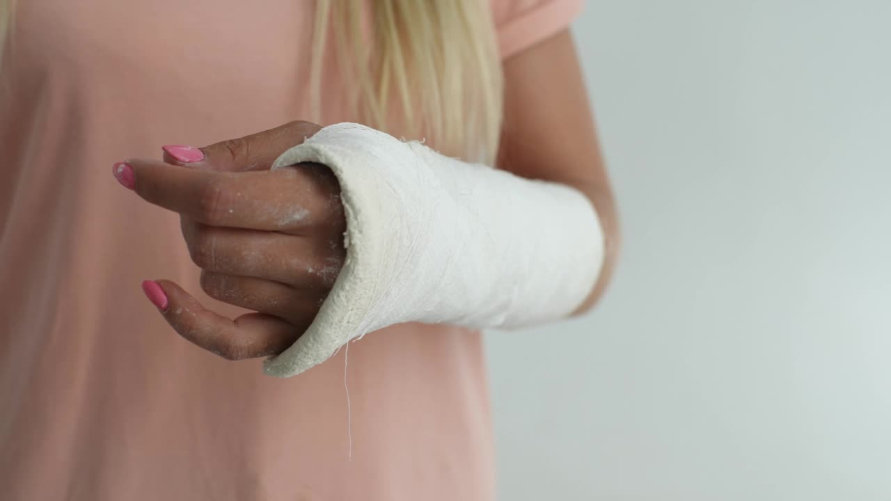 特写裁剪的镜头，无法辨认的年轻女子破碎的手臂包裹在白色石膏绷带轻轻按摩手腕在白色孤立的背景。视频素材