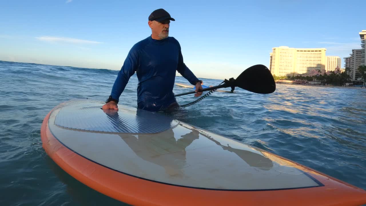 在夏威夷瓦胡岛檀香山的威基基，一名男子带着SUP站起来在橙色冲浪板上冲浪。视频下载