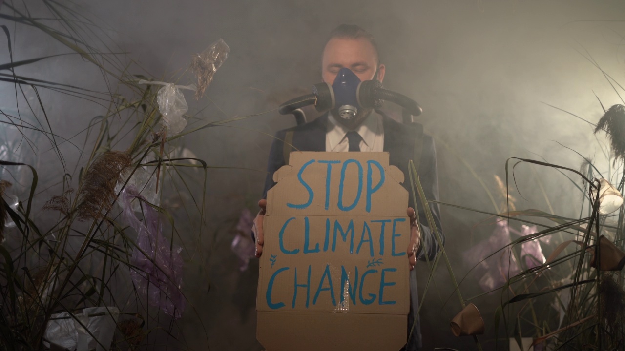 一名留着胡子的白人男子戴着氧气面罩，带着充气气球，站在满是塑料垃圾的干草中间，举着“停止改变气候”的牌子。生态灾难的概念视频素材