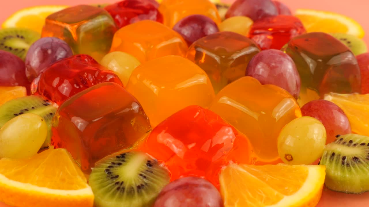 多颜色的浆果明胶果冻与葡萄，橘子片和猕猴桃在粉红色的背景。用勺子敲击后晃动的果冻。视频下载