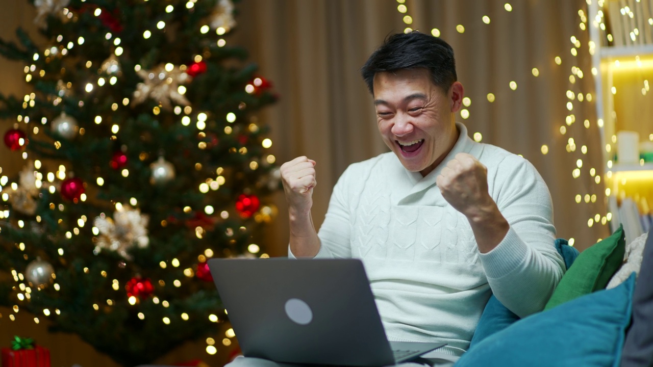 兴奋的亚洲人喜出望外地庆祝伟大的网络新闻，坐在沙发上看着笔记本电脑屏幕享受成功，享受商业优惠，出售折扣，互联网彩票胜利，赢视频素材