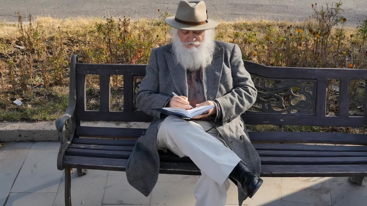 穿着大衣、帽子、裤子和锃亮的鞋子，留着白色蓬松胡子的时髦优雅的高级男子坐在户外的长凳上，用圆珠笔在笔记本上做笔记。年老的商人视频下载