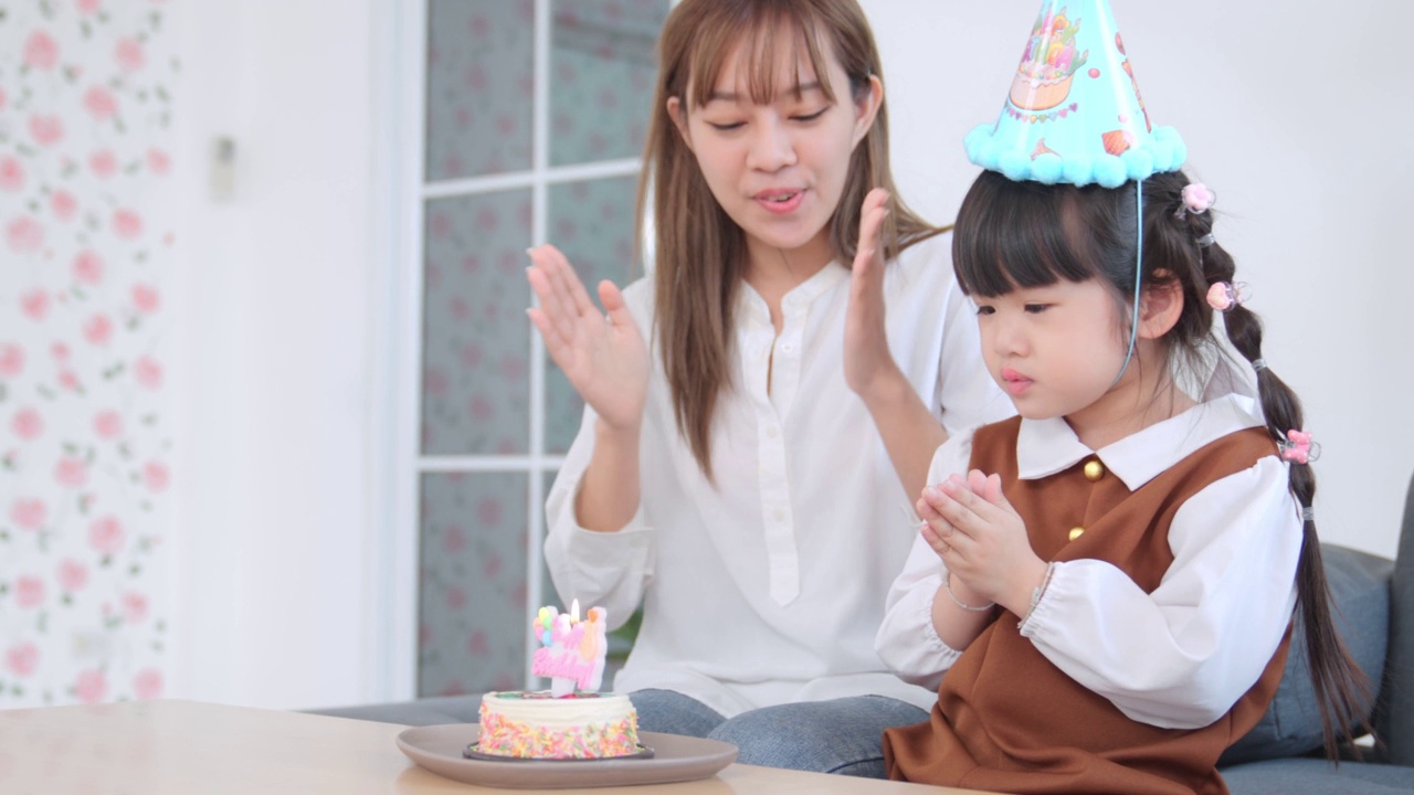 和妈妈在客厅为小女孩的生日聚会，亚洲孩子的女孩吹灭生日蛋糕蜡烛，亚洲妈妈庆祝女儿的生日视频素材