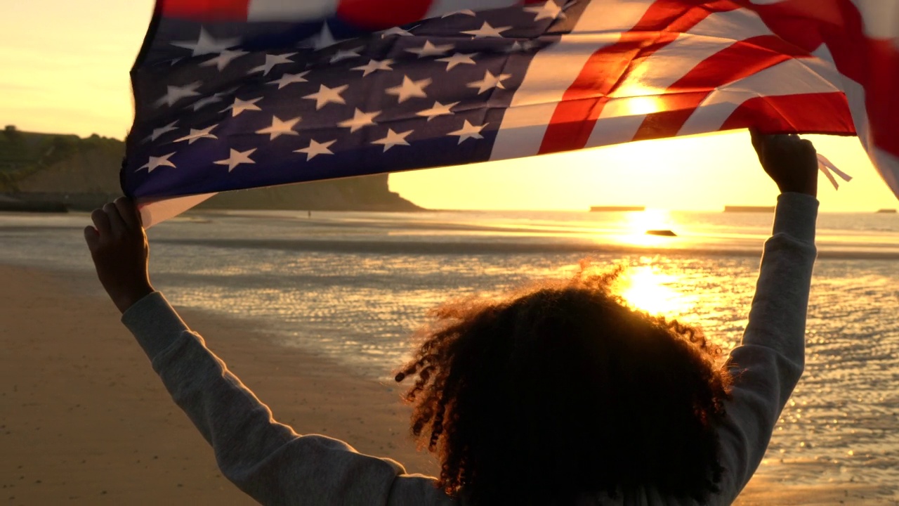 混合种族，混血，非裔美国女孩，青少年，女性，年轻的女人在日落或日出的海滩上拿着美国星条旗视频素材