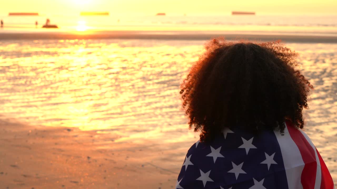 非裔美国女孩，十几岁的女性，年轻的女人裹着美国星条旗，看着人们在日落或日出的海滩上玩耍视频素材