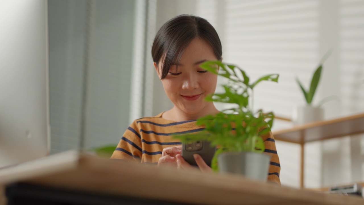 亚洲妇女坐在家里办公室快乐地微笑着拍了一张植物的照片并分享到社交媒体上。视频下载
