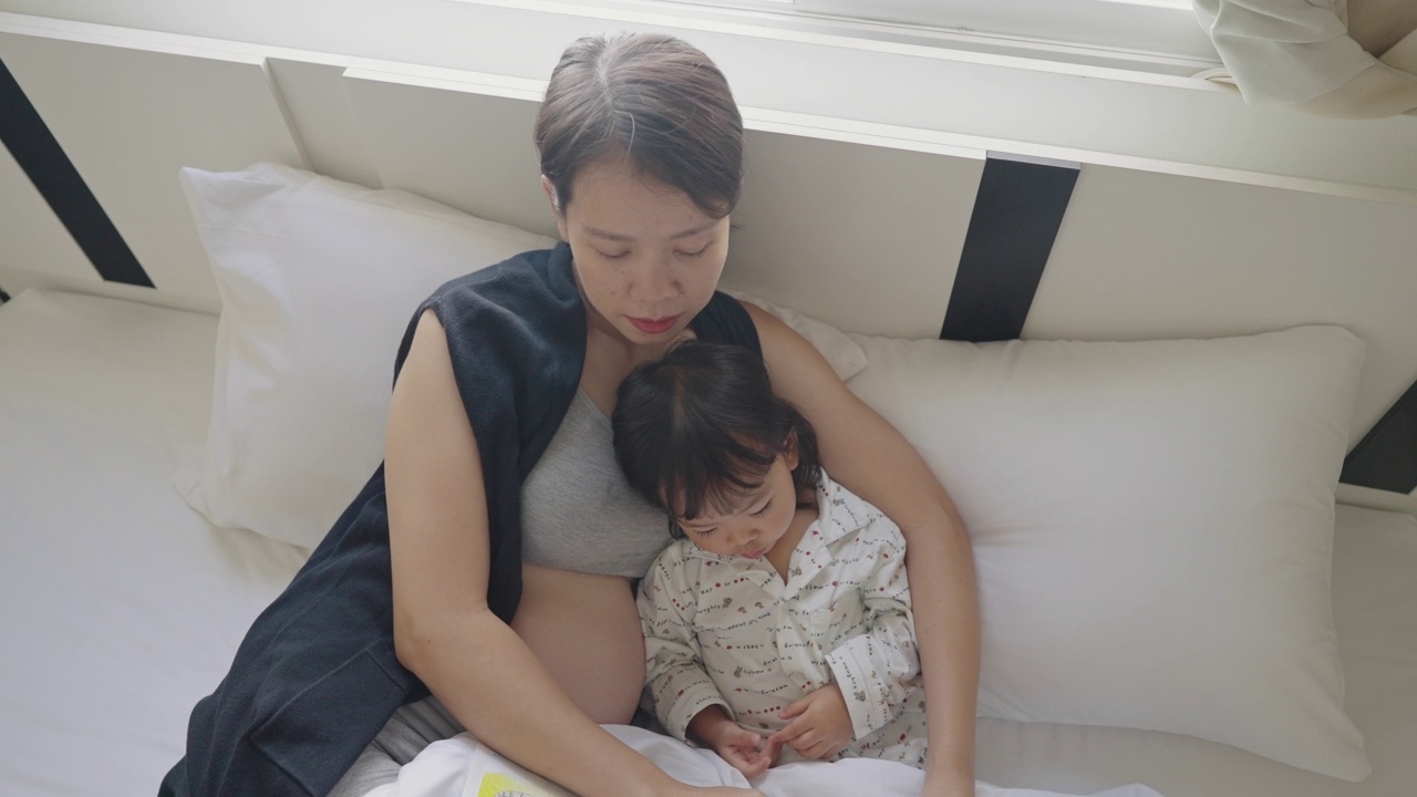 一个亚洲小孩在看绘本，她的妈妈也怀孕了，早上躺在床上视频下载