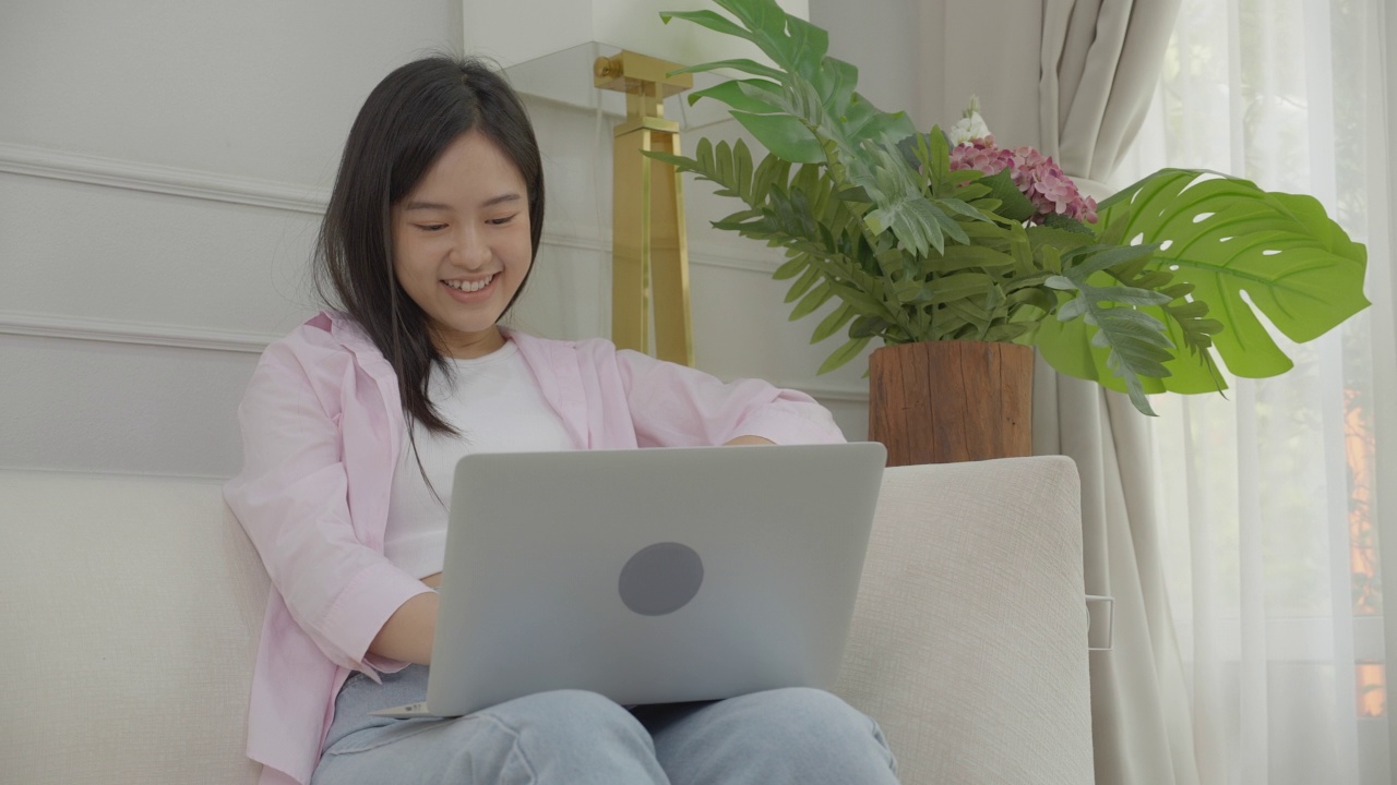 幸福年轻的亚洲女性坐在沙发上，在笔记本电脑上工作，而成功和兴奋在家里，自由职业女性使用笔记本和成就，商业和生活方式的概念。视频素材