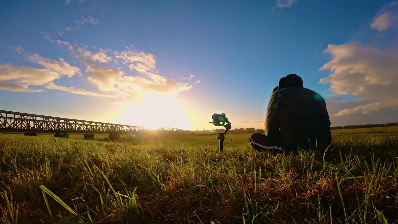 男子摄影师与相机在万向架拍摄迷人的时间在铁路附近与云在天空日落。从早到晚视频下载
