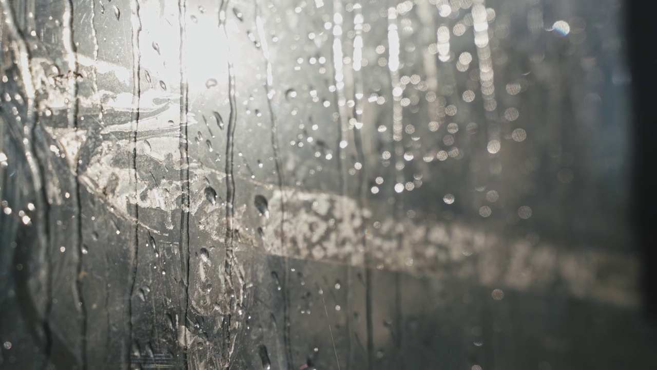 从玻璃上滴下的雨滴特写。小雨滴顺着玻璃流下，映衬着阳光明媚的早晨。水滴从玻璃上流下来。视频素材