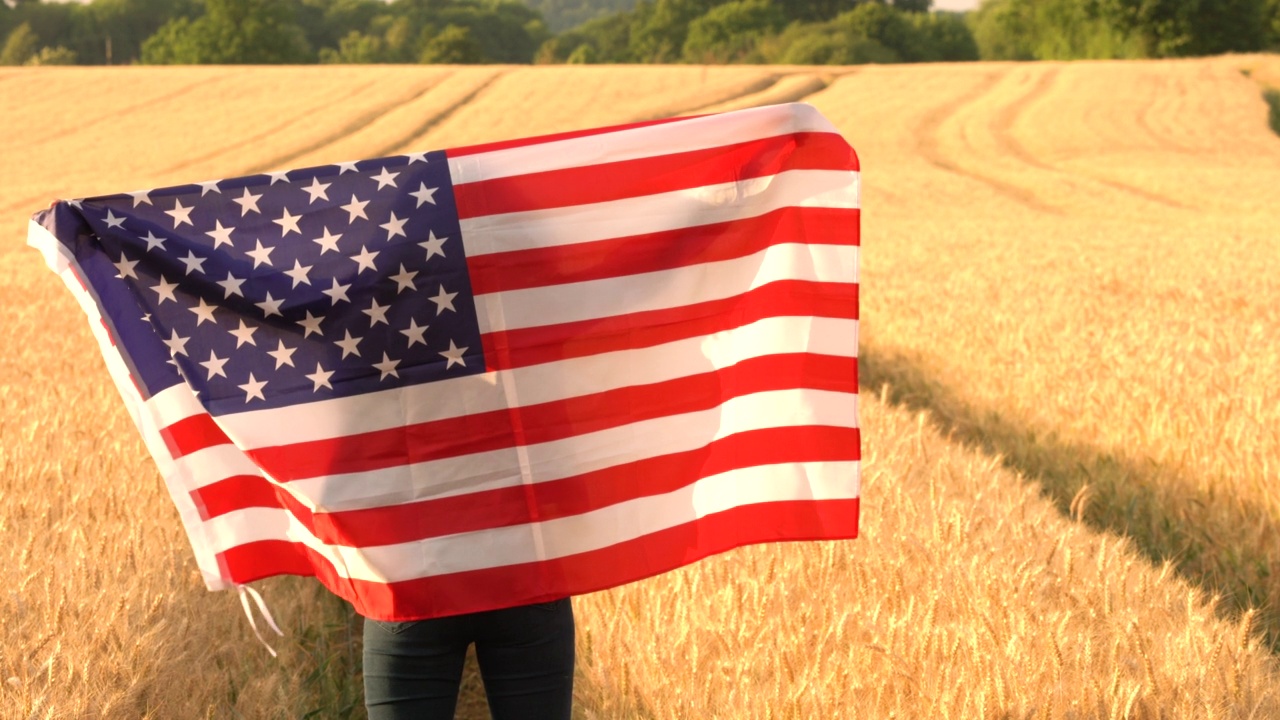 美国女孩，十几岁的女性，年轻的女人在她的头顶麦田里拿着美国星条旗，然后在日落或日出时把它裹在她的身上视频素材