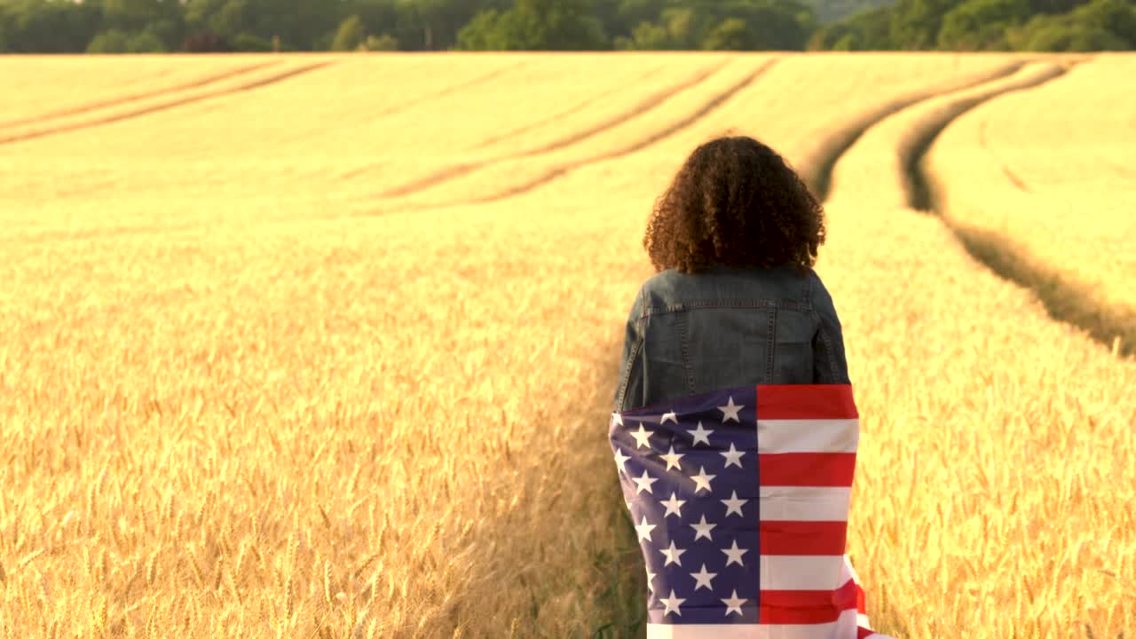 一个非裔美国女孩，十几岁的女性，年轻的女人，裹着美国星条旗，在日落或日出的麦田里把它举过头顶视频素材