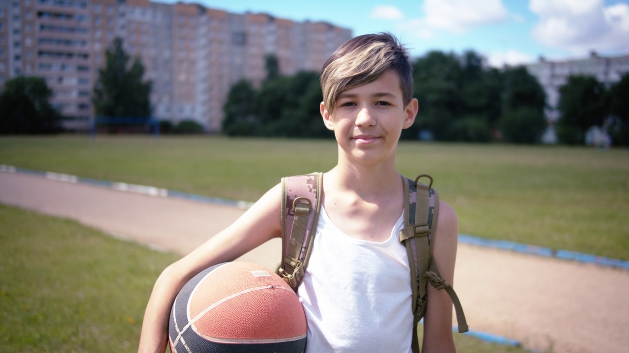 十几岁的男孩带着一个篮子球看着相机微笑，在夏天阳光明媚的日子，移动相机视频下载