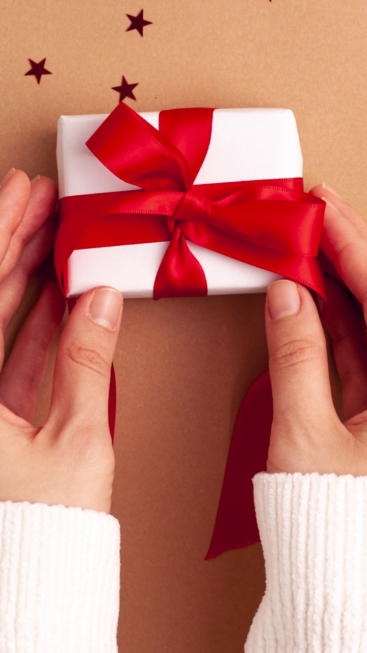 穿着白色毛衣的人手拿走了一个白色的纸礼物，红色缎带蝴蝶结在棕色的背景和红色的星星形状。定格动画垂直圣诞假期概念平铺视频下载