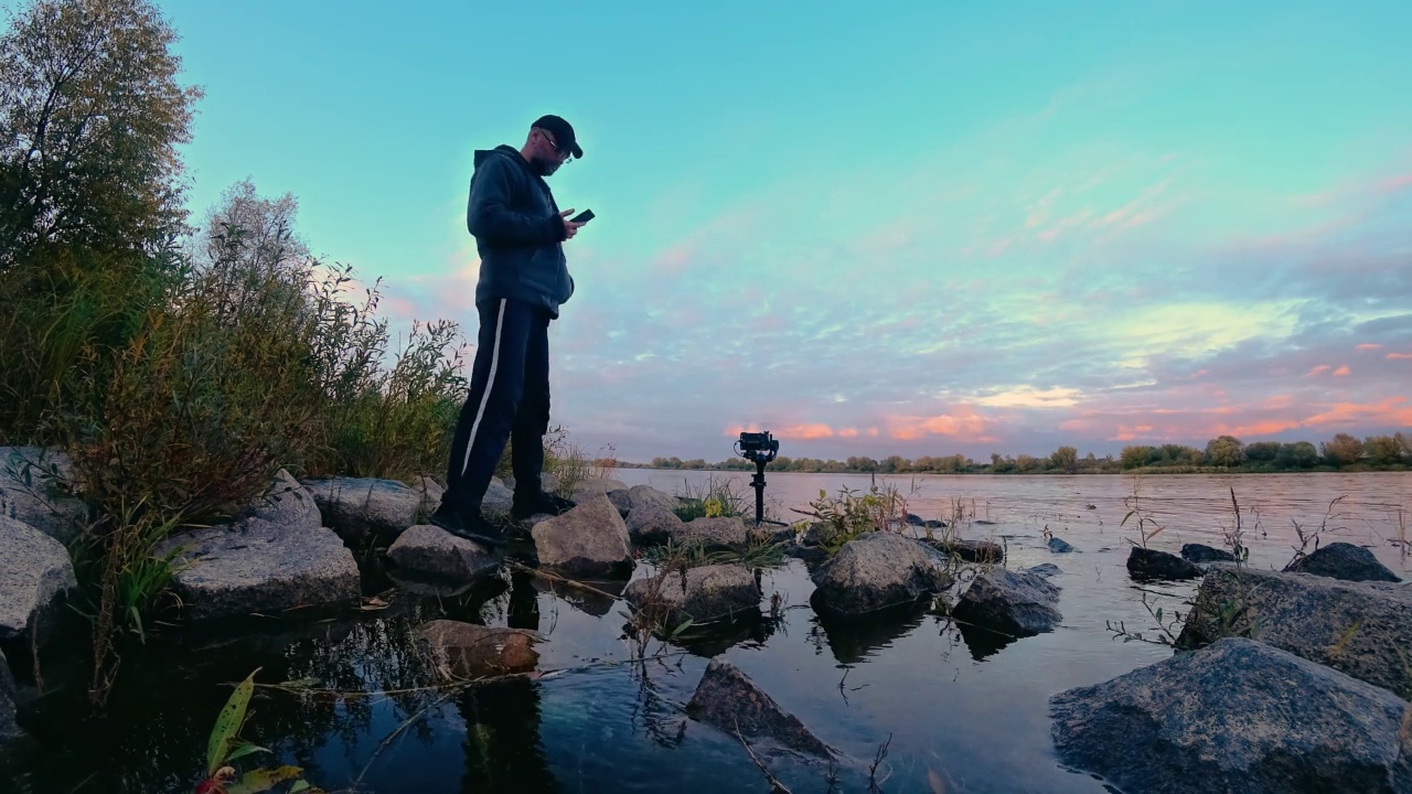 男子摄影师与相机在万向架拍摄时间流逝的云在天空与河岸的倒影与水中的石头视频素材