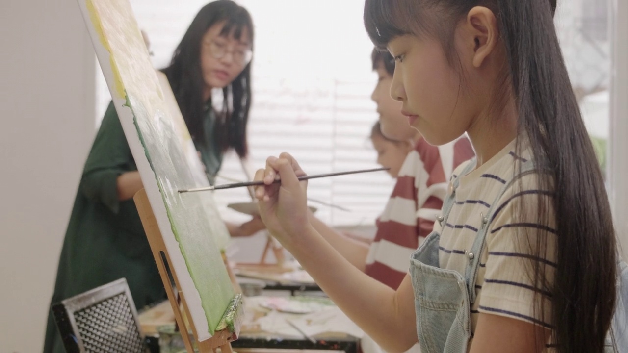 在美术教室里，一个小女孩正专注地在画布上画亚克力彩画。视频素材
