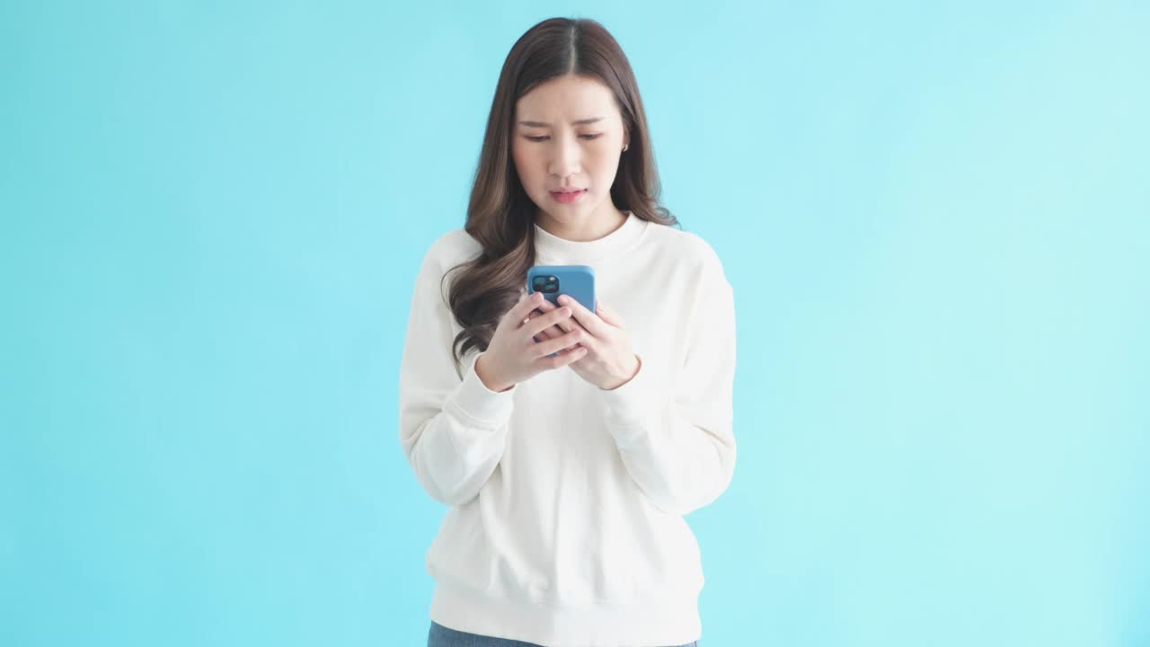 年轻的亚洲女子在手机短信中读到坏消息。站在孤立的蓝色背景下，女性悲伤地看着智能手机视频素材