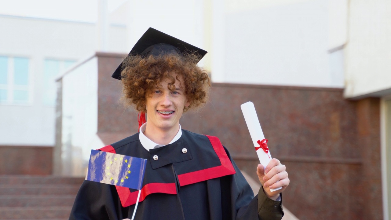 一个卷毛的、滑稽的研究生披着硕士斗篷，手里拿着毕业证书和欧盟国旗视频下载