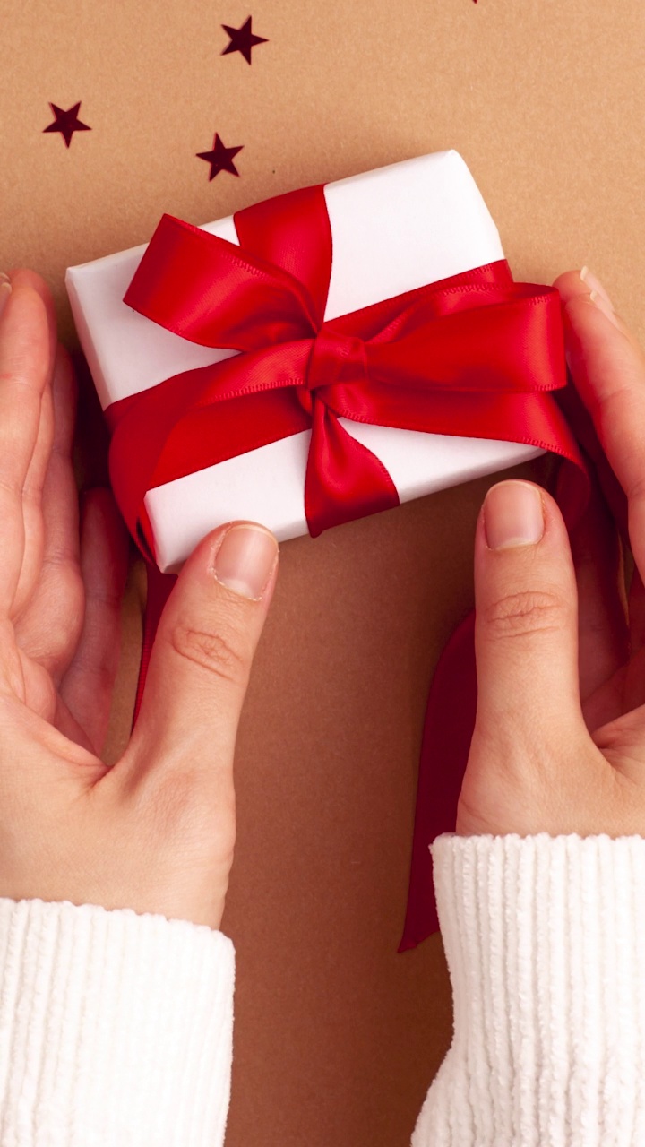 人的手在白色的毛衣拿着一个白色的纸礼物，红色缎带蝴蝶结在棕色的背景与红色的星星形状。定格动画垂直圣诞假期概念平铺视频素材