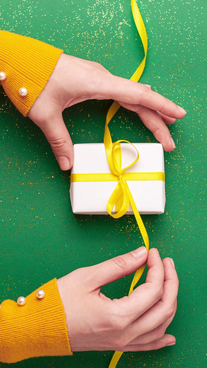 人类的双手松开，绑了一个黄色缎带蝴蝶结白色礼品盒玉绿色背景与金色闪光。定格动画垂直圣诞假期和现在平铺视频下载