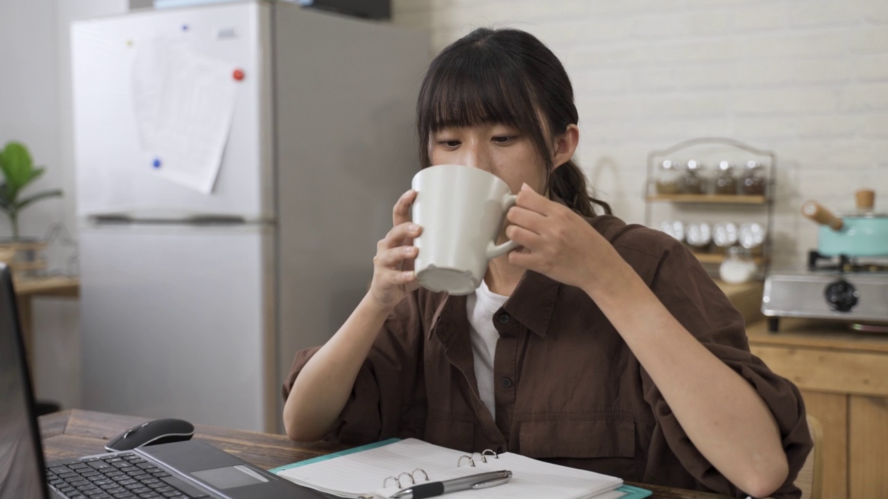 一位在电脑前工作的亚洲妇女在明亮的室内，坐在餐桌边喝着美味的咖啡，微笑着叹息视频素材