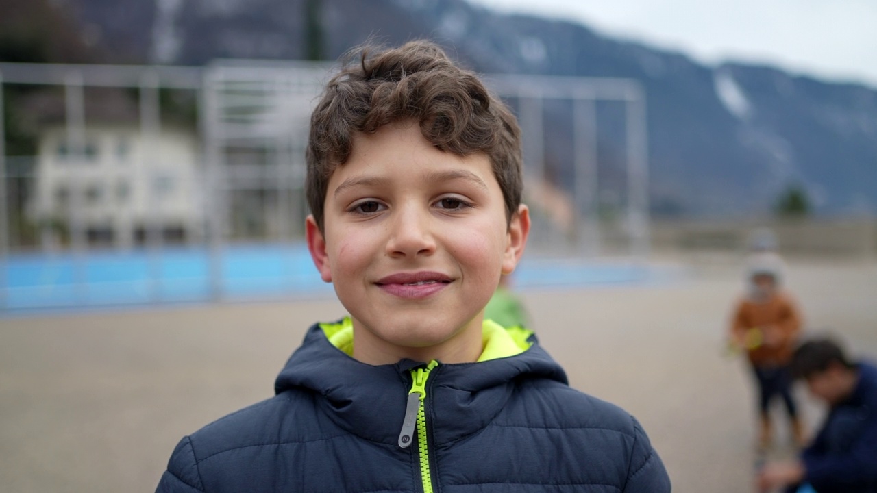 一个英俊的小男孩站在城市公园外对着镜头微笑。男性前十几岁的孩子穿冬天的衣服夹克的肖像脸特写。快乐的情绪视频下载