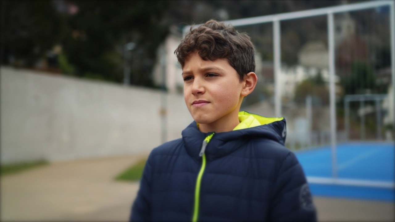一个深思熟虑的小男孩站在城市公园外面，表情严肃。英俊的十几岁前的男孩穿着冬装在沉思视频素材