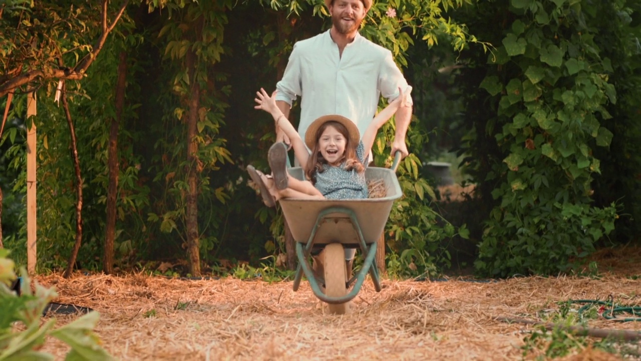 戴着草帽、满脸胡须的微笑男子推着一辆手推车，旁边是一个快乐的小女孩。父亲和女儿一起在花园里享受夏天的周末。缓慢的运动。园艺的概念视频下载