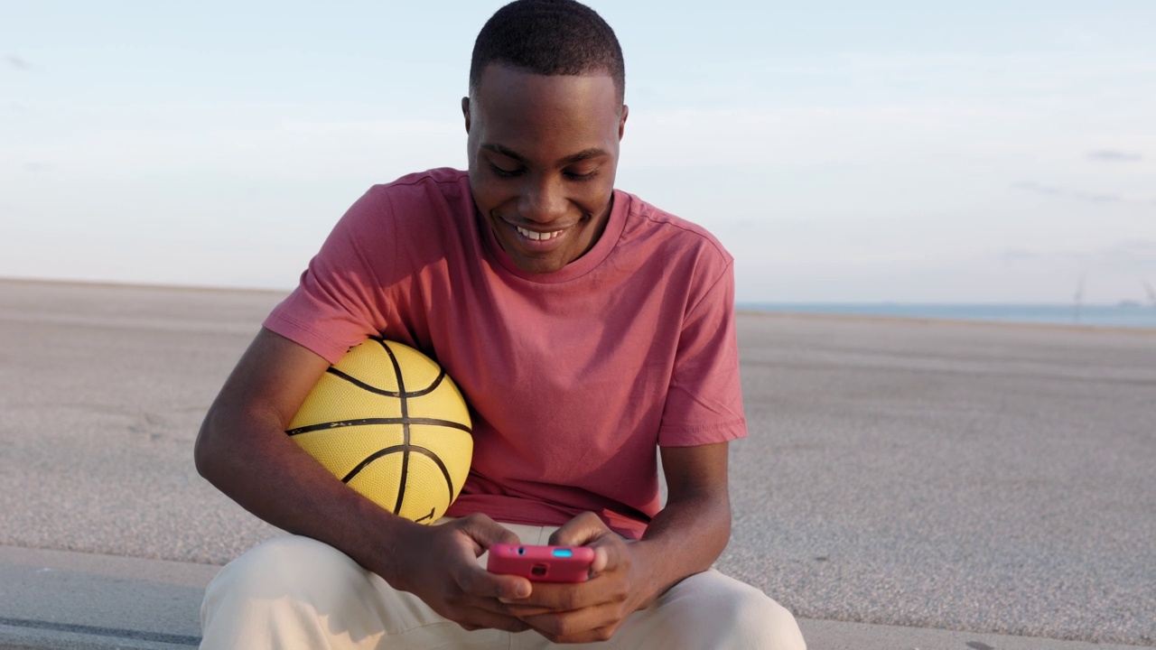 年轻的非裔美国人穿着t恤坐在户外使用智能手机视频下载