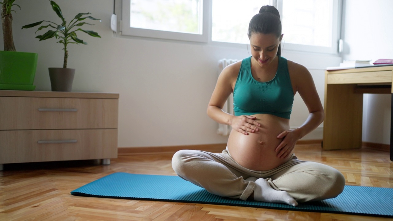 孕妇，揉着她的腹部，坐在莲花位，在家里练习瑜伽视频素材