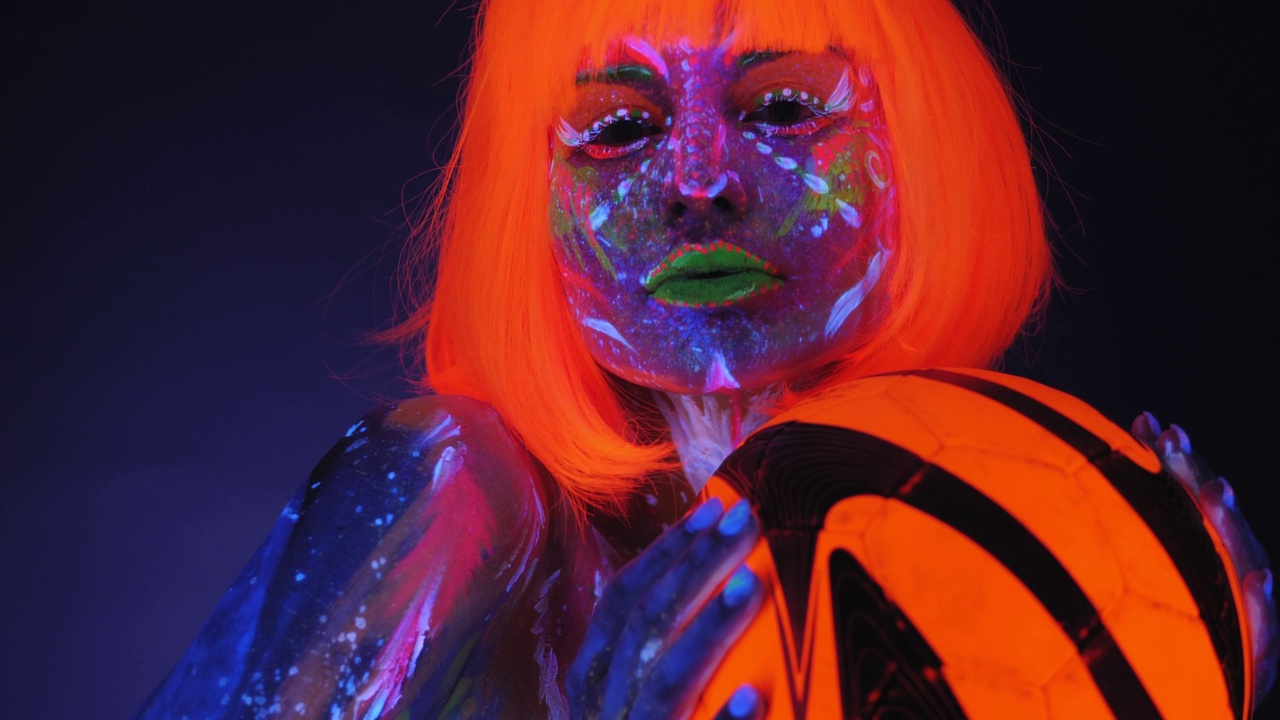 一名戴着亮橙色假发的紫外线人体艺术女子手持一个橙色球。视频下载
