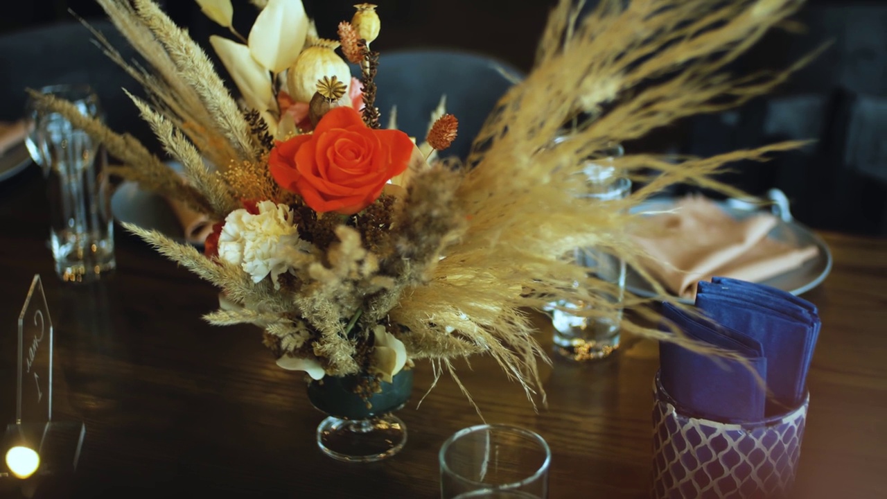 装饰婚礼的餐桌装饰有鲜花、红玫瑰和潘帕斯草，玻璃杯和盘子，相机移动的慢动作。视频素材
