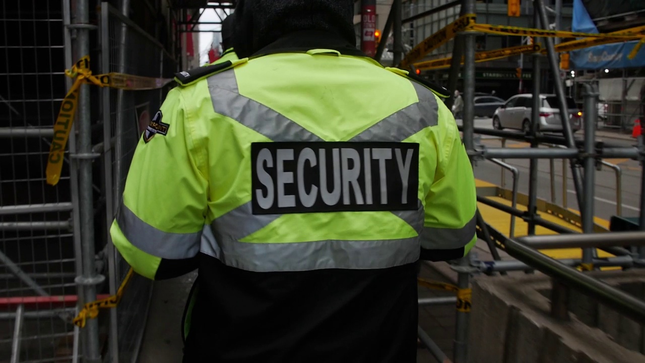 高清图中穿着霓虹绿色安全外套的保安人员在金属脚手架之间行走的后视图视频下载