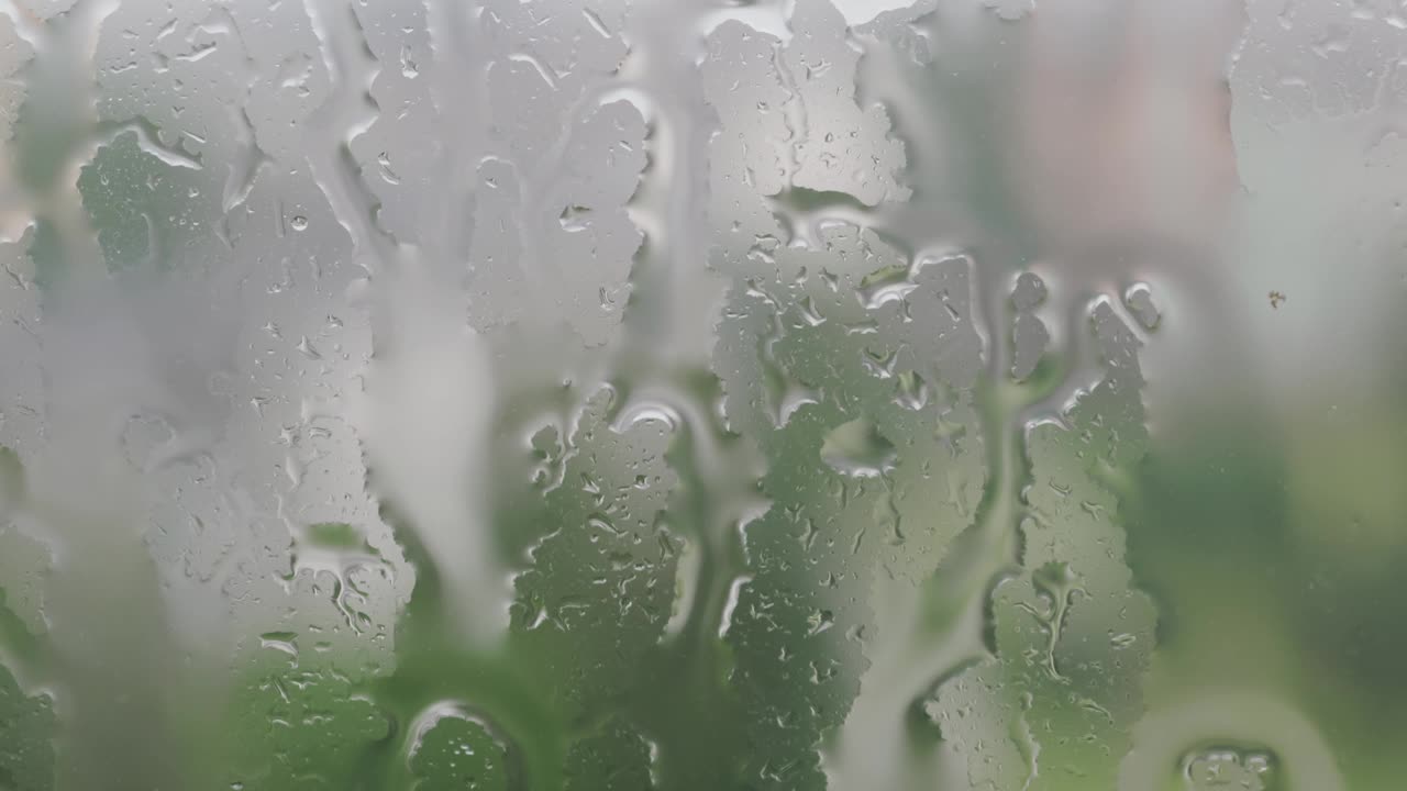 特写雨点水滴在大雨中滴落在潮湿的窗户玻璃上，背景与树木的剪影模糊视频下载