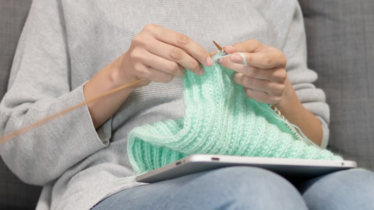 女人把平板电脑放在腿上，看着屏幕，织着毛衣。妇女学习编织，观看视频教程，在线课程，遵循编织模式视频下载