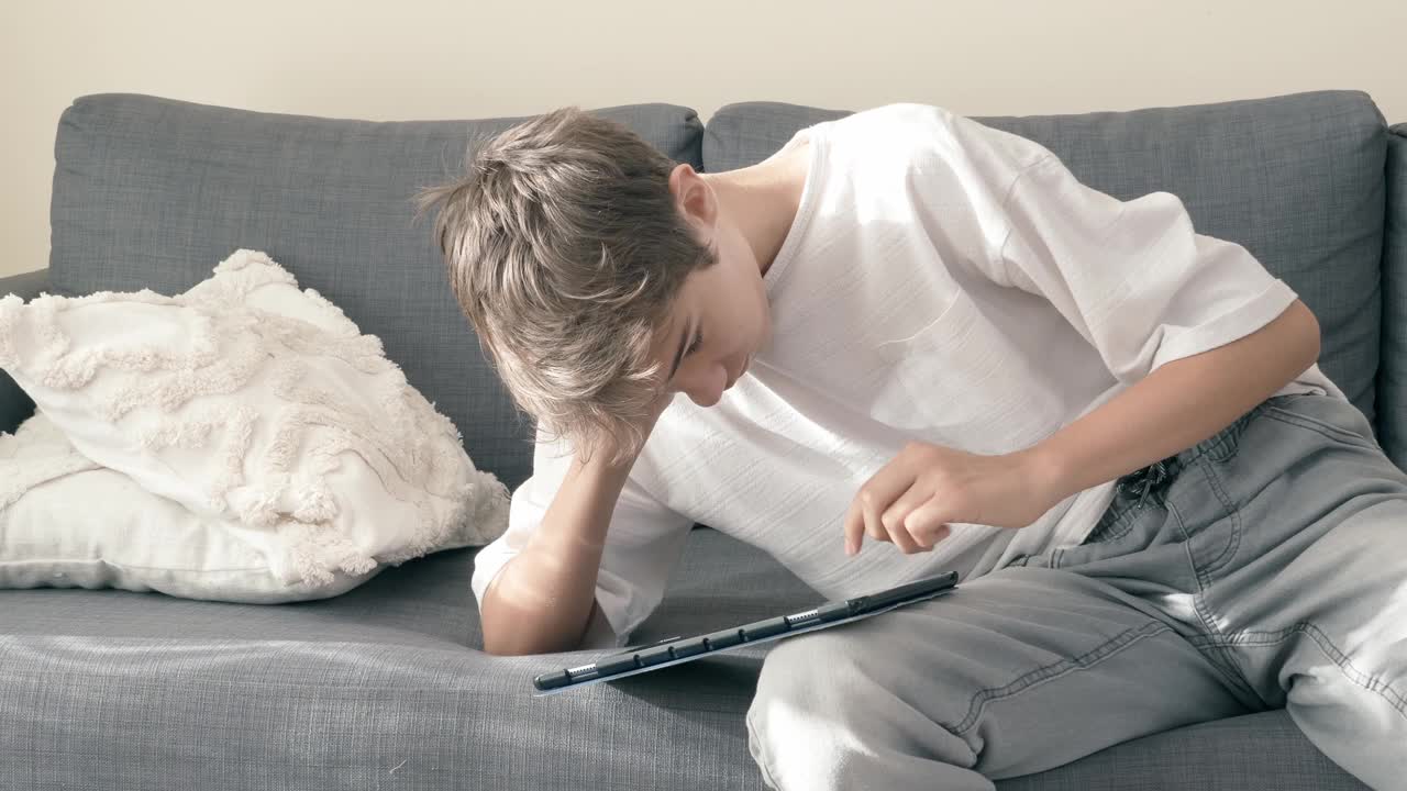 十几岁的男孩在家里的沙发上使用数字平板电脑。孩子看着小工具显示，触摸屏幕，滚动，使用应用程序玩在线视频游戏，浏览互联网，看视频视频下载