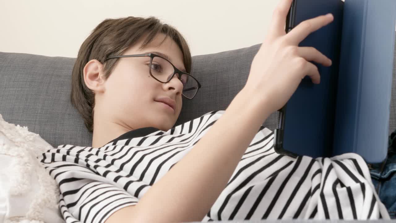 十几岁的男孩在家里的沙发上使用数字平板电脑。孩子看着小工具显示器，触摸屏幕，使用应用程序玩在线视频游戏，浏览互联网，看视频视频素材