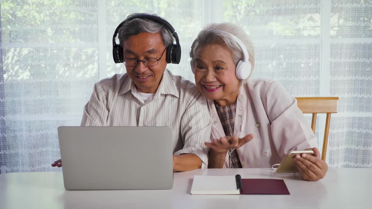 快乐的亚洲老夫妇一边在家里玩笔记本电脑和手机，一边享受着听音乐视频素材