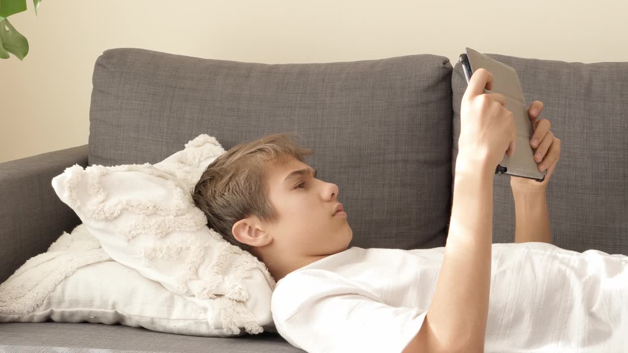 青少年在家中使用数码平板电脑。男孩在平板电脑上玩视频赛车游戏，躺在家里的沙发上视频下载