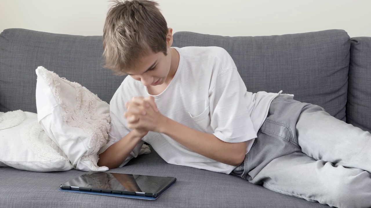 十几岁的男孩躺在家里的沙发上使用平板电脑。快乐的孩子享受成功，得到好消息，赢得比赛，通过考试在学校视频素材