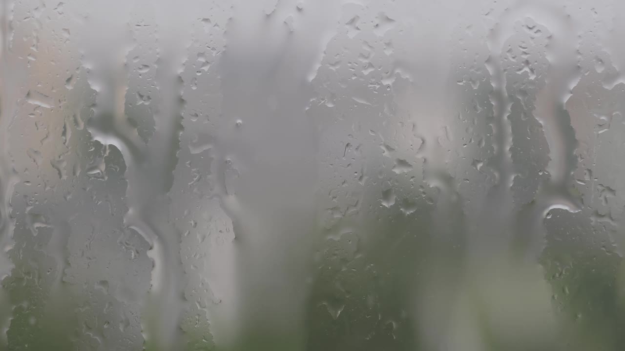 特写雨滴水滴在大雨中滴落在潮湿的窗户玻璃上，背景模糊，树木的剪影视频下载