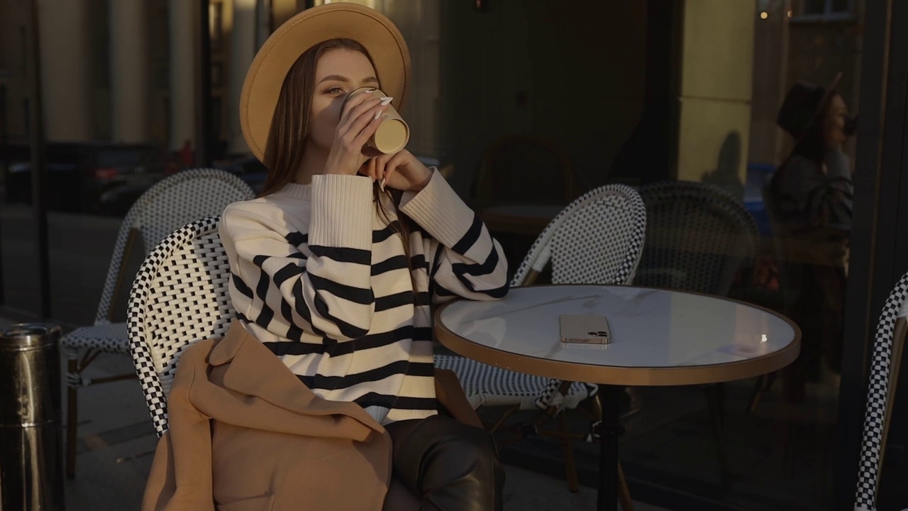 年轻漂亮的女孩戴着帽子坐在街上的咖啡馆里喝着早茶或咖啡。在露台上享受咖啡视频素材