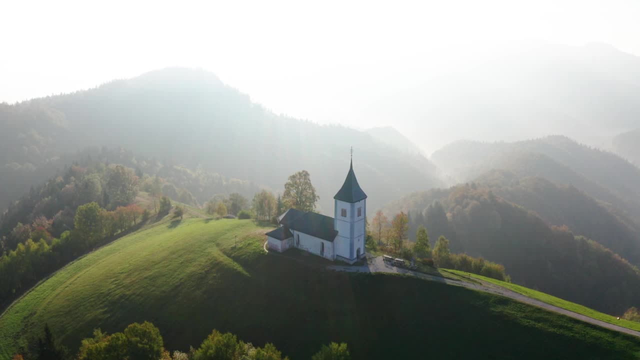 鸟瞰山上的小教堂和秋天日出时的低云。斯洛文尼亚。鸟瞰雾中山上美丽的教堂，绿色的草地，树木，秋天黎明的天空。景观视频素材