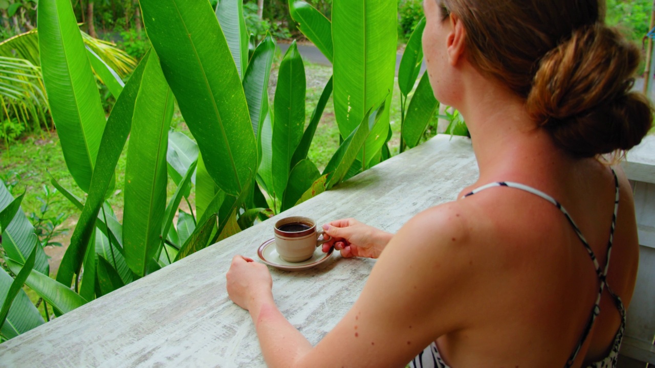 一名女子闻到并尝试着在早晨喝热腾腾的巴厘岛咖啡，享受绿色的热带花园自然。特写后方的孤独的旅行女孩在巴厘岛努萨佩尼达印度尼西亚。视频下载
