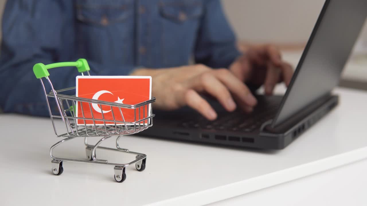 桌上有土耳其国旗的小购物车。男子用笔记本电脑在网上购物。视频下载